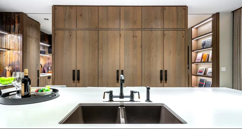 A dual-basin sink in a run of floor-to-ceiling wood panel cabinetry with an illuminated bookshelf.